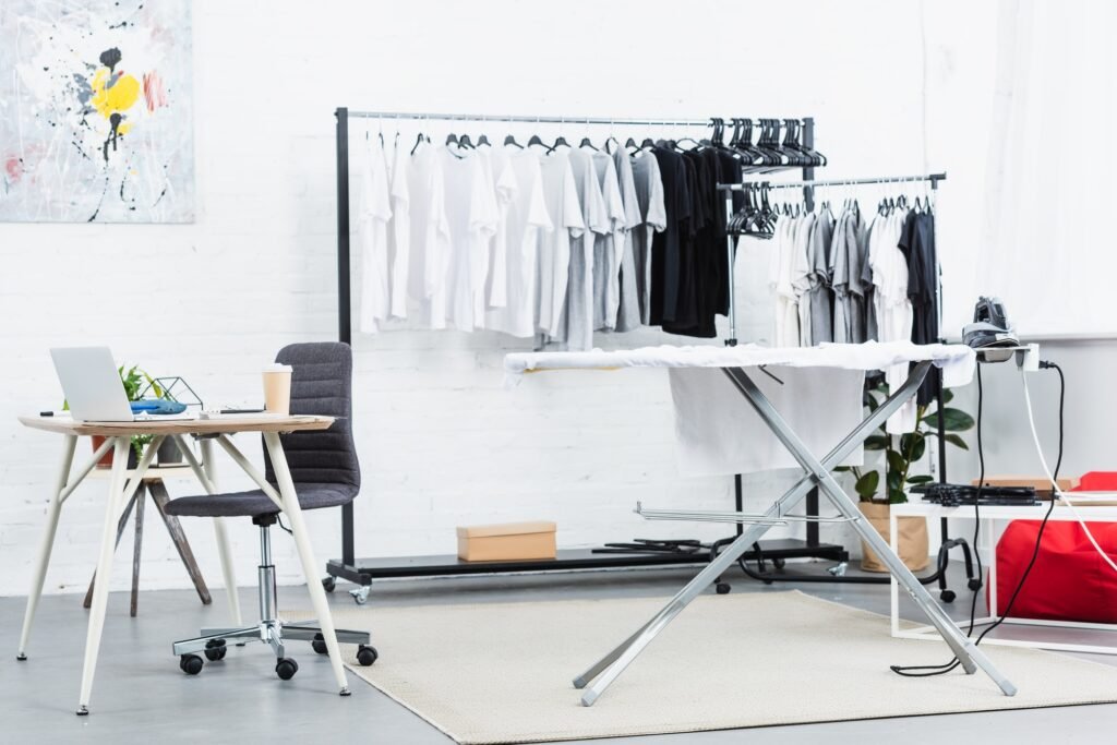 selective focus of t-shirts on hangers, table with laptop and ironing board in clothing design